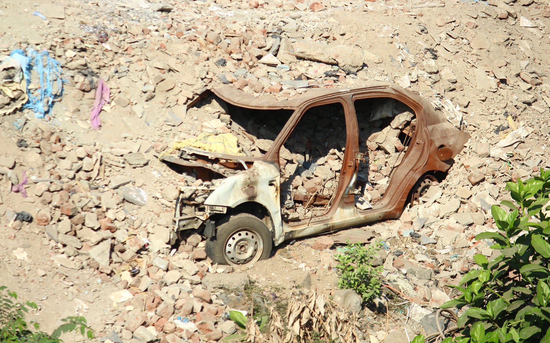 A rusted, broken car shell partially buried in rubble and dry earth, surrounded by debris and scattered plants. The scene captures a sense of abandonment, decay, and the passage of time.